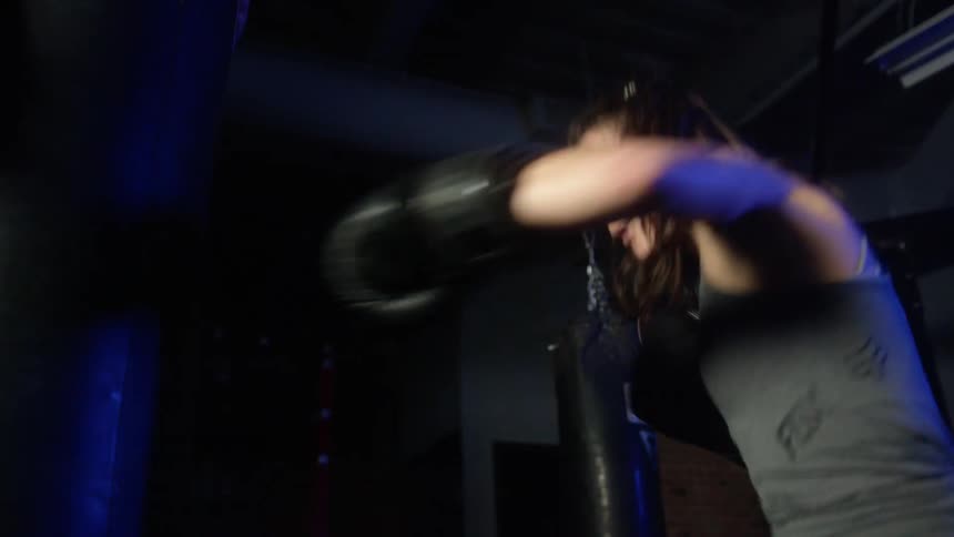 Female boxer with pony tail and boxing gloves hits heavy bag in boxing gym