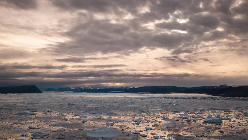 A lake with water and ice with a mountain range the sky and clouds in the background