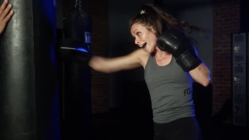 A woman in a gym with boxing gloves punching a heavy bag as a man spots
