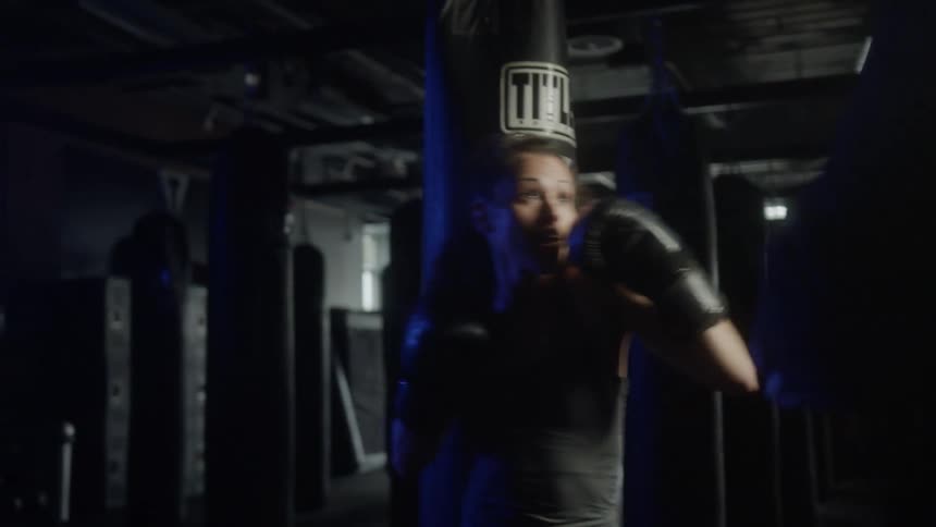 A woman in a gym with boxing gloves practicing boxing with heavy bags in the background