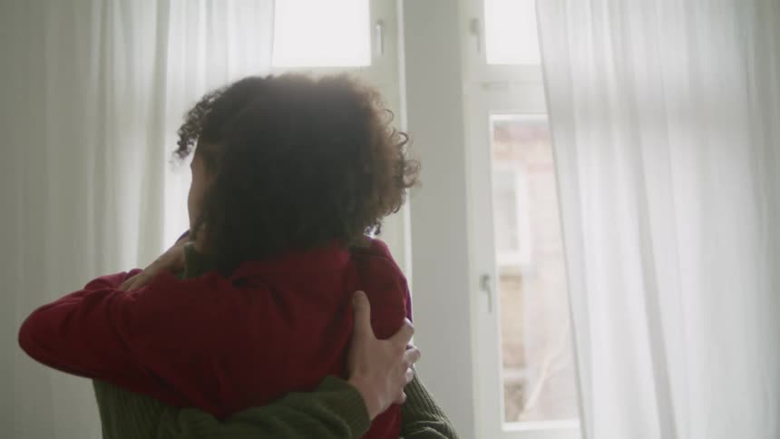 Man with green sweater and woman with frizzy hair and red sweater lovingly cuddle each other in room