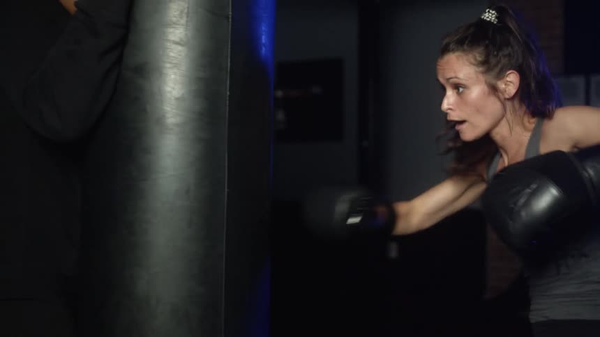 A woman with boxing gloves punching a heavy bag as a man spots it