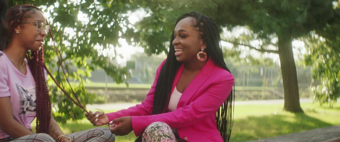 Woman sitting in park strokes another womans braids then her own