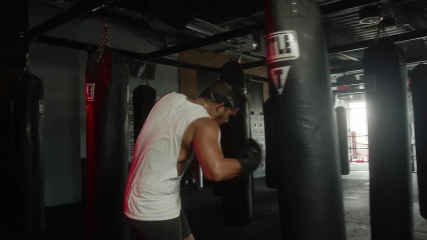 A man wearing black boxing gloves punching a black punching bag in a room with a bunch of other black punching bags