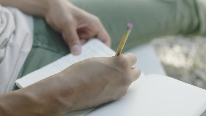 Man holding a journal in his lap writing something down with a pencil at daytime