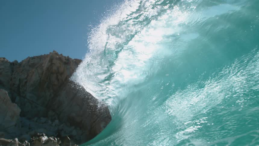 The sun shines on a big blue wave splashing as it rolls overhead near rock formations