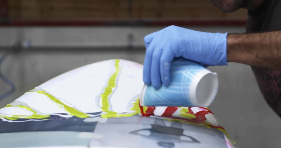 A man with a glove pouring paint from a cup on a surfboard with a wall in the background