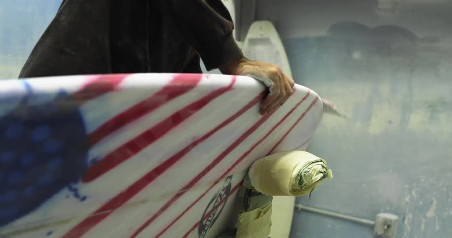 A man wearing a respirator mask hand sands a painted surfboard