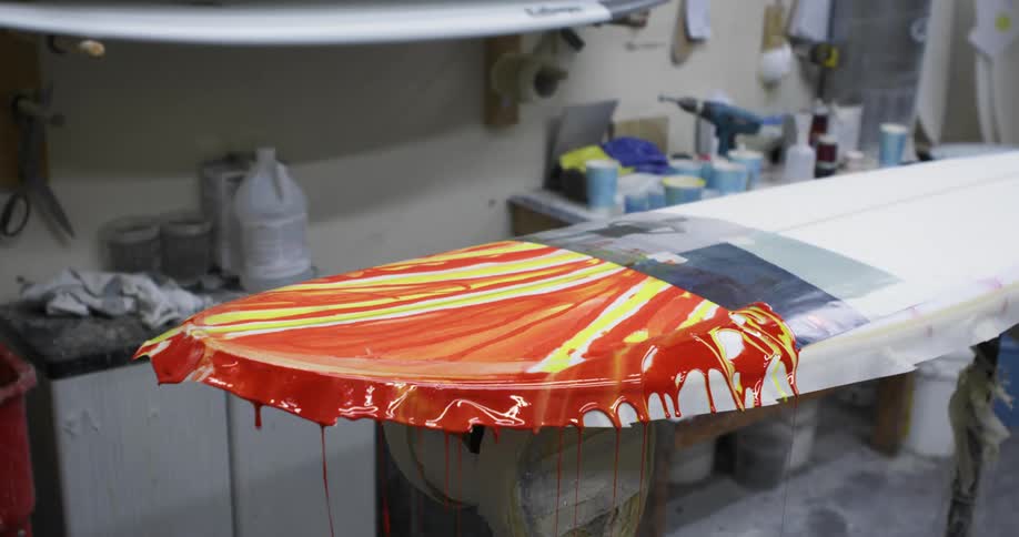 A man spreads orange and yellow paint on a surfboard in a scraping motion