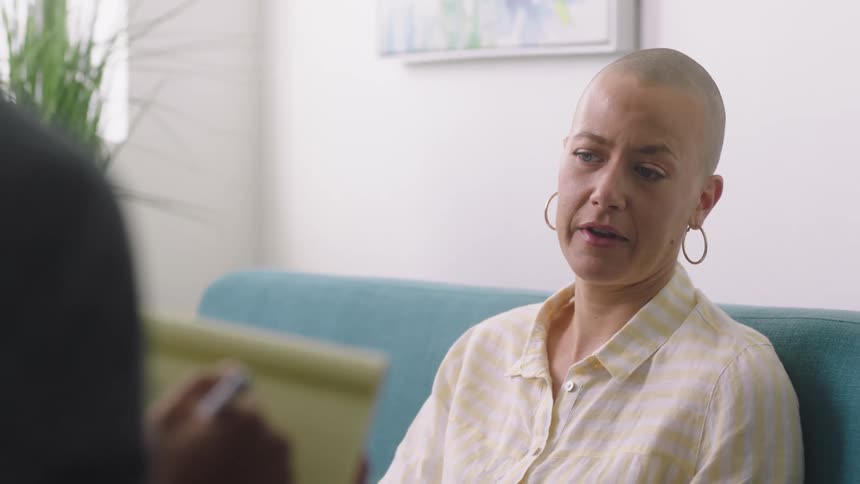 Bald woman with blue eyes wearing metallic earrings talking to someone taking notes in a room