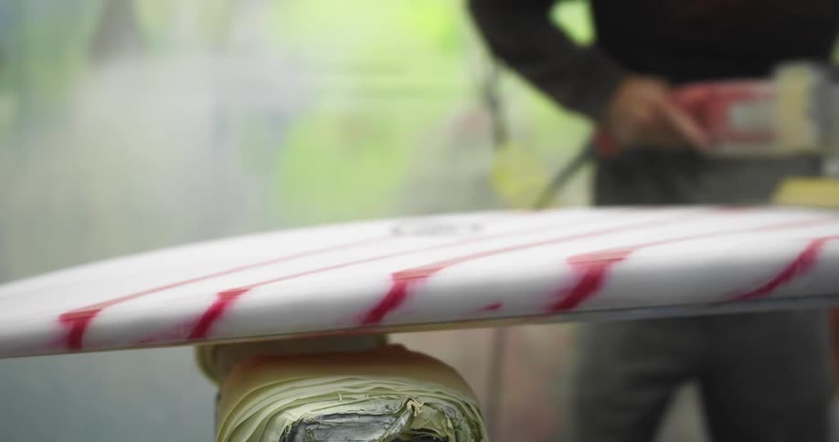 A man sands down the top of his white and red striped surfboard in his workshop