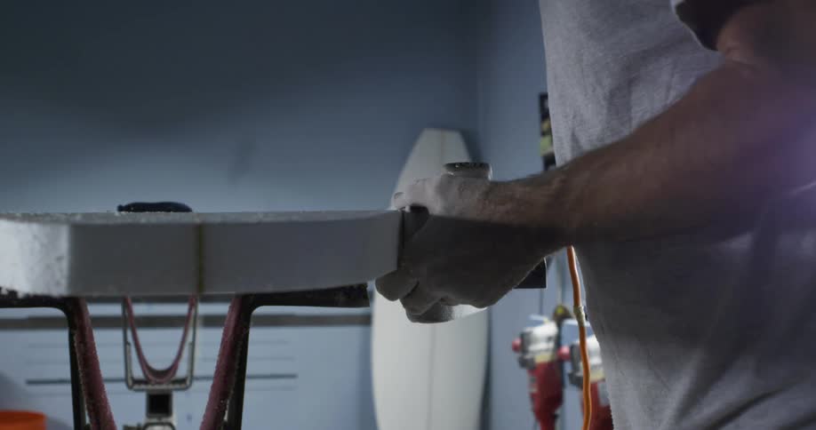 A person uses a power tool to sand the edges of a surfboard inside a workshop