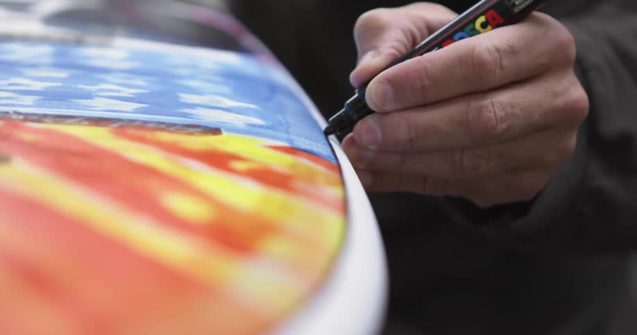 A man drawing a line with a black marker over the surface of a surfboard inside a workshop