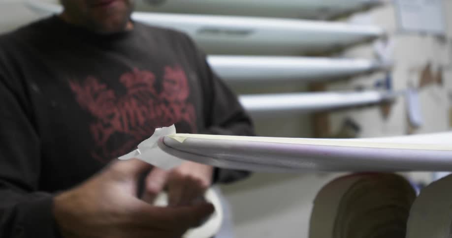 A man covers part of a surfboard in tape in a repair shop