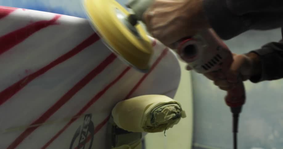 A man with a mask sanding a surfboard with an electric sander