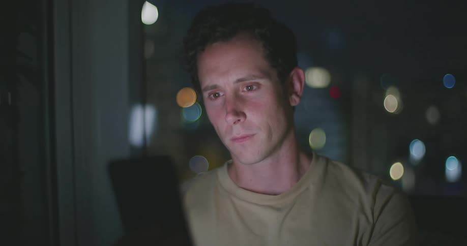 A man holds and taps on his phone with city lights glowing through the window behind him