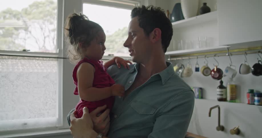 A man holding a small girl in his arms attempts to cheer her up while standing in the kitchen