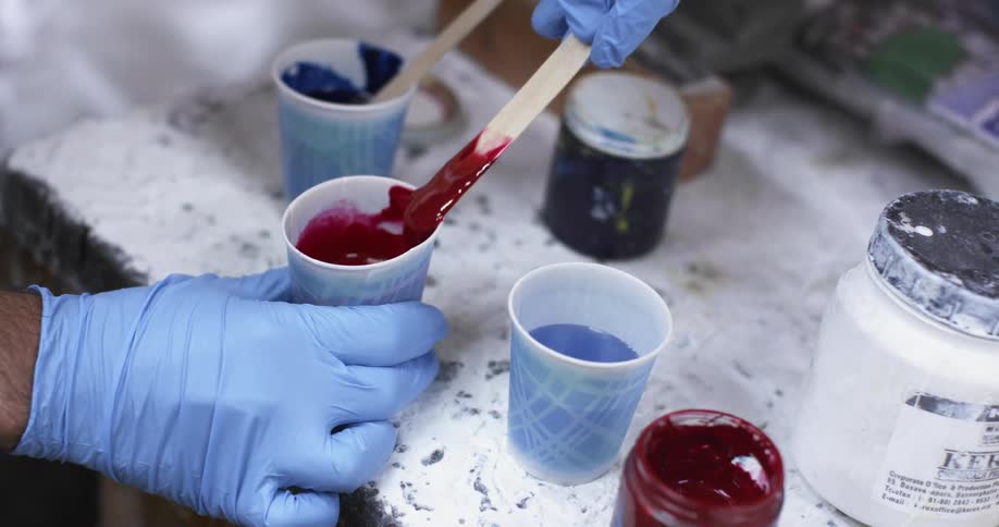 A person wearing latex gloves stirring red paint in a small cup