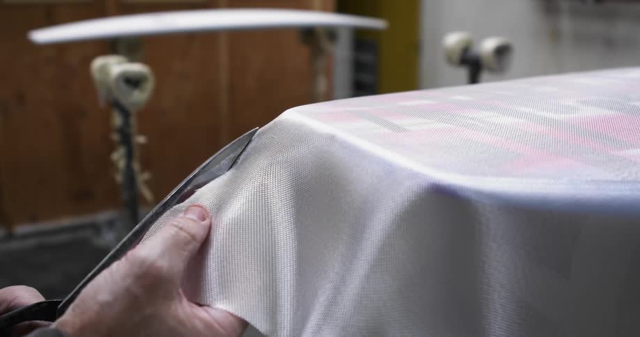 A man uses scissors to cut fabric on the surface of a surfboard in a workshop