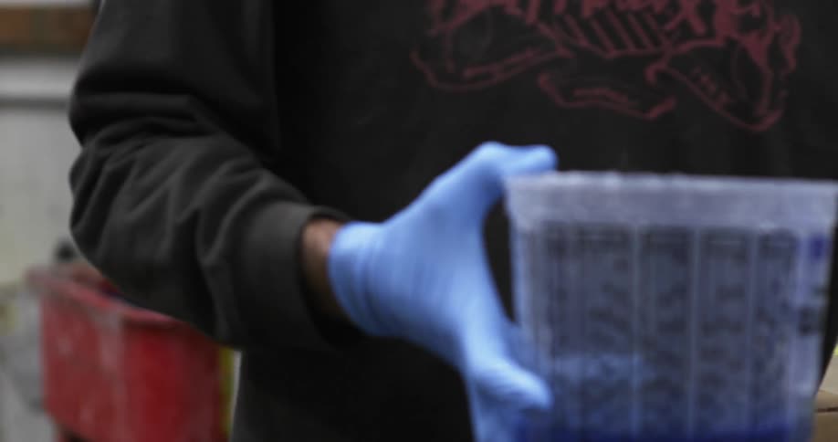 A man wearing gloves pours a liquid into a container he is holding then stirs it