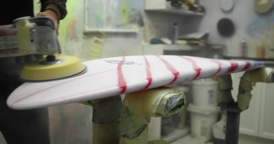 A man in a workshop using a buffering tool to smooth the surface of a unfinished surfboard