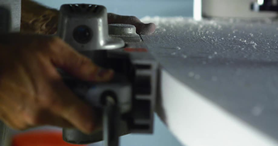 A person uses a power tool to sand the edges of a surfboard inside a workshop