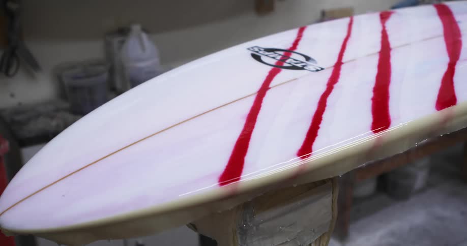 A man walks into his workshop where a surfboard sits partially painted with red stripes