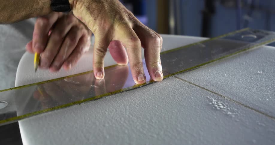 A person in a room marking a surfboard with a pencil and ruler