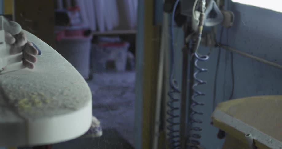 A person uses a power tool to sand the edges of a surfboard inside a workshop