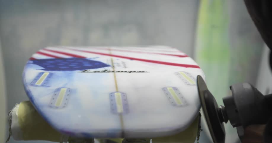 A person uses a spinning buffer machine to smooth the sides of a surfboard on a crafting workbench