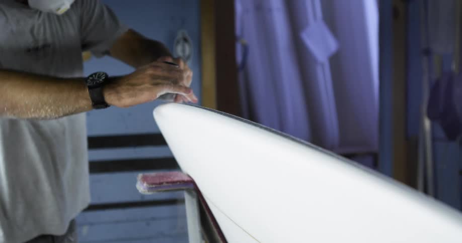 A man with a mask and a wristwatch sanding a surfboard