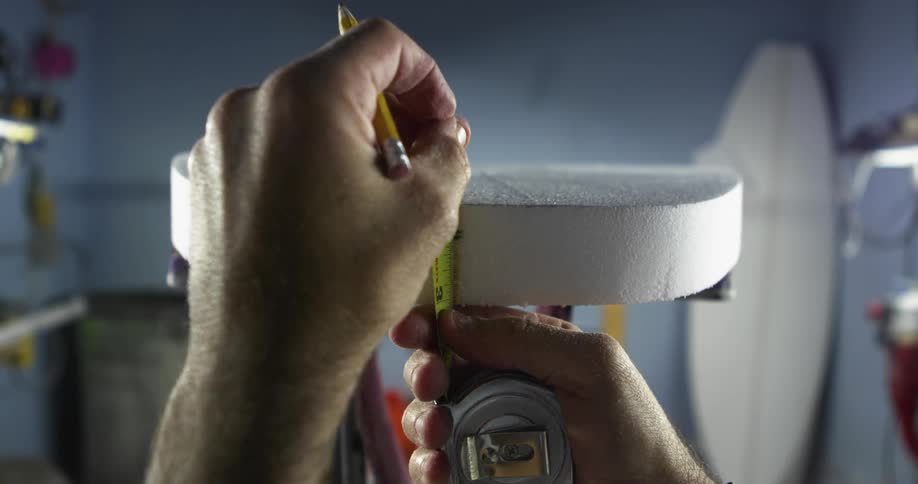 A person uses a pencil and a measuring tape on the edge of a surfboard