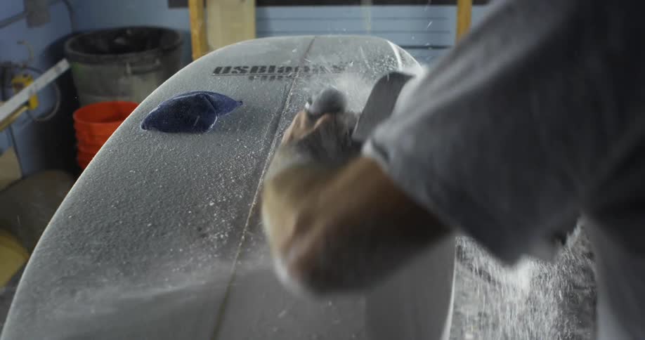 A man sanding the surface of a surfboard with a power tool in a workshop