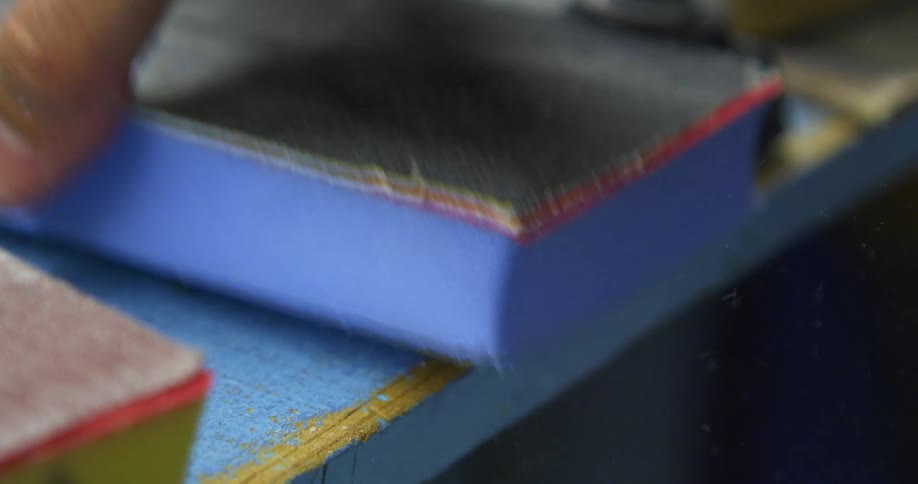A hand places a square blue object on the surface of a blue and yellow piece of wood