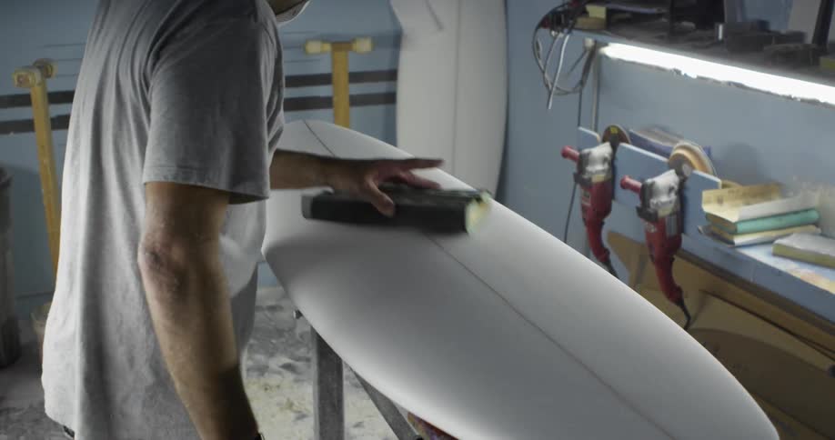 A man sanding the surface of a surfboard in a workshop