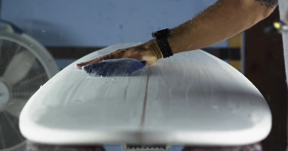 A man sanding a surfboard cleans the debris using compressed air