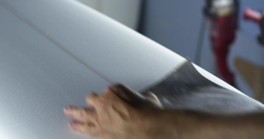 A man sanding the surface of a surfboard by hand in a workshop