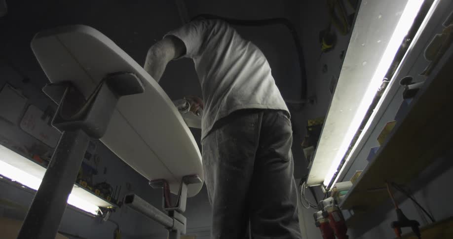 A man sanding a surfboard with a power tool in a workshop