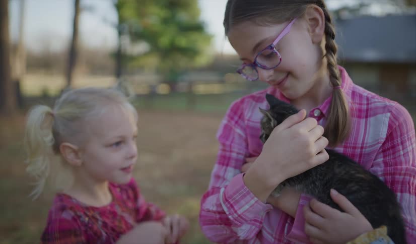 An older sister holds a small kitten in her arms while her younger sister plays with it with a piece of string outside in a yard during the day