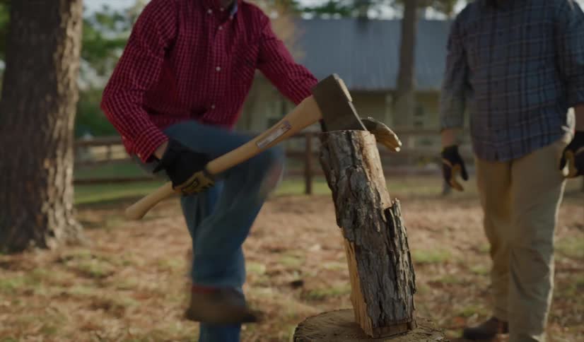 Father standing next to his son who swings an ax down from over his head and hacks at a piece of wood on a tree stump outdoors during the day