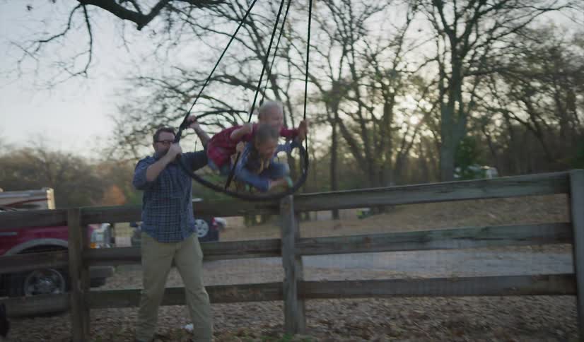 A man pushing two children on a swing with trees the sky and clouds in the background