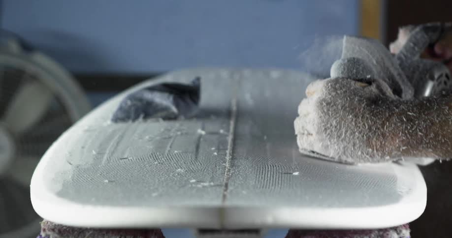 Dust flies as a man sands a surfboard