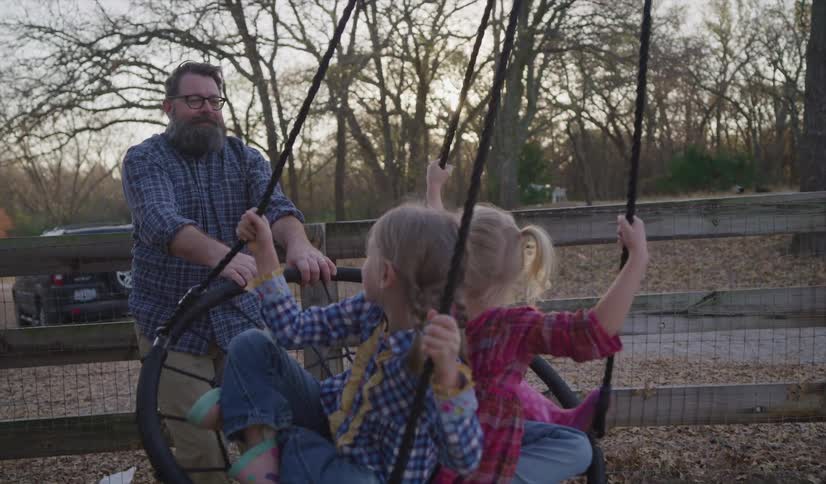A man smiles as he pulls back a tire swing that his daughters are seated in at a park
