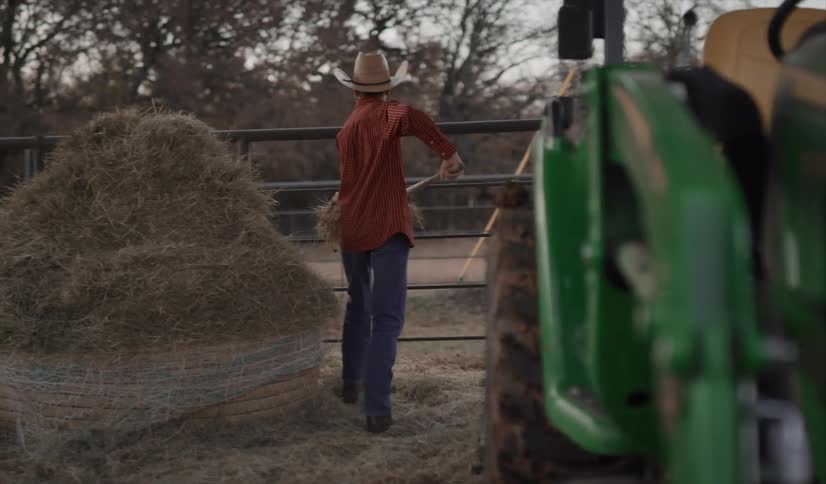 A boy with a hat moving hay with a pitchfork next to a tractor with trees the sky and clouds in the background