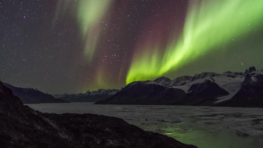 Aurora borealis shining brightly in a star filled sky above snow covered mountains and ice covered lake