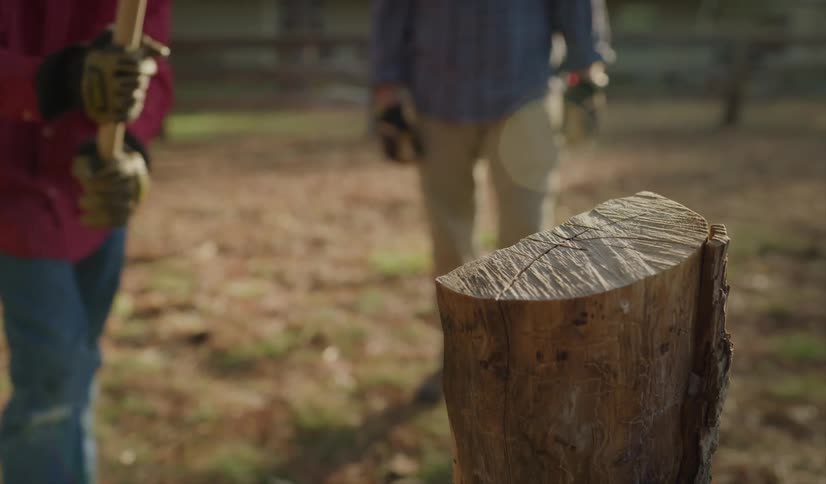 A boy swings an ax and struggle to split a log as his father watches