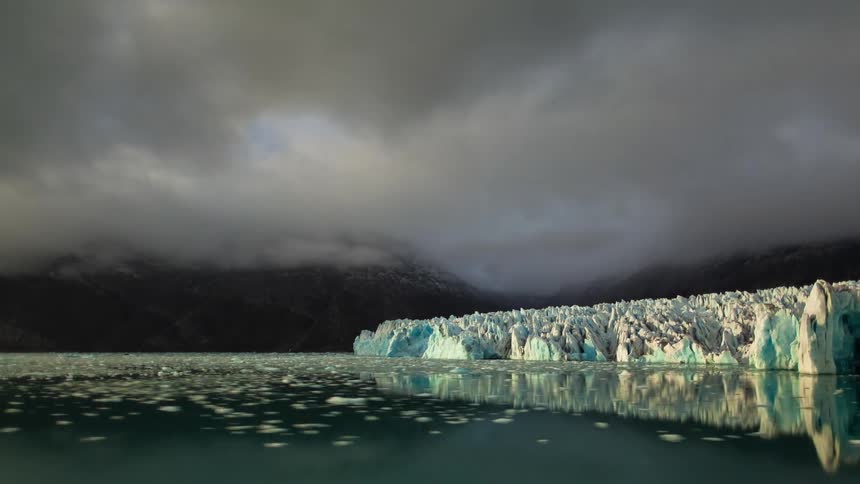 Time lapse of an arctic lake next to a rocky terrain with pointed glaciers and low mist moving overhead and northern lights appearing at night