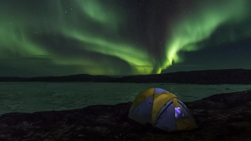 Tent alongside an ice covered lake and the aurora borealis is shining brightly in a star filled sky