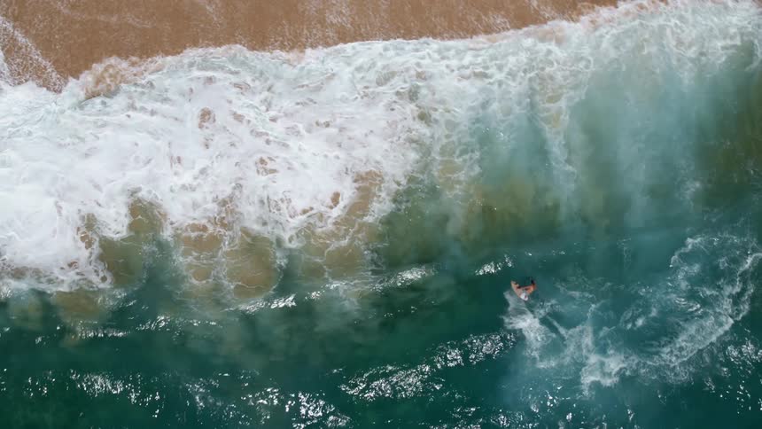 Aerial view of a man riding an ocean wave and crashing into the shore of a beach