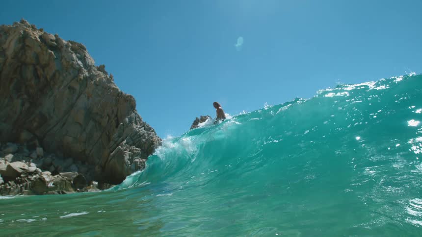 Surfer rides a big blue wave near rock formations under a clear blue sky